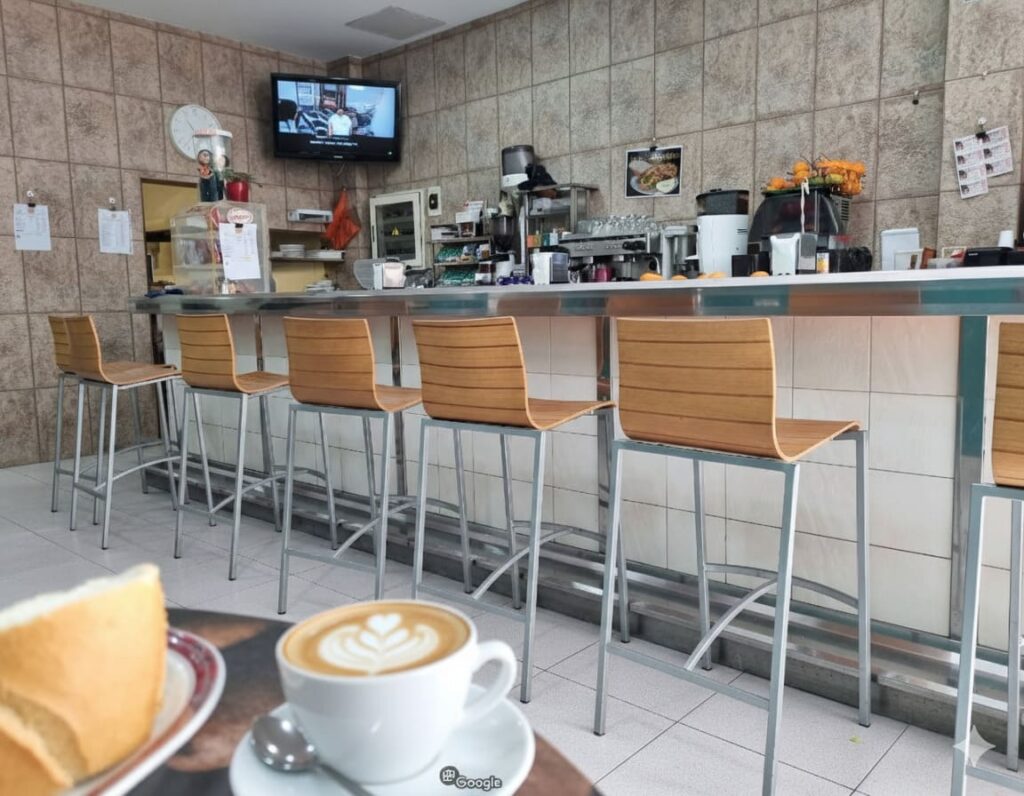 Interior de la "Cafetería del 15" en Puerto del Rosario con una barra larga y seis taburetes de madera, una televisión montada en la pared y una taza de café con arte latte en primer plano.
