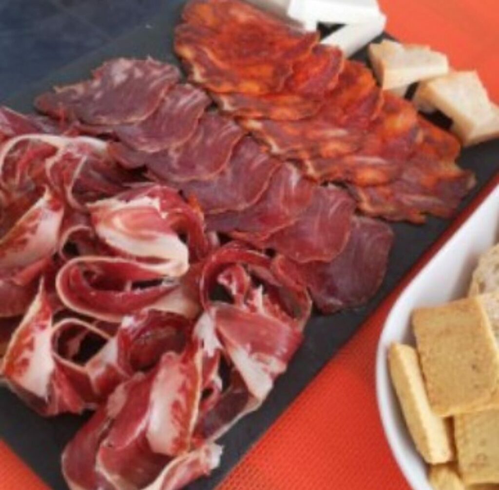 Tabla de embutidos ibéricos variados, incluyendo jamón, lomo y chorizo, servida en una pizarra junto a trozos de queso y picos de pan.