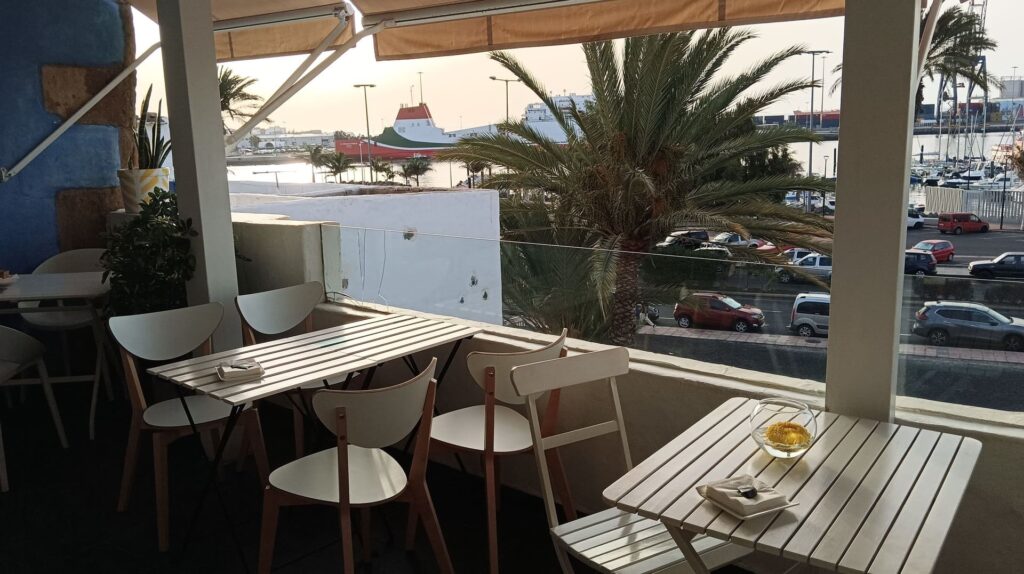 Terraza del restaurante Zielo Terraza al atardecer, con mesas y sillas blancas, y vistas al puerto con un ferry y palmeras.