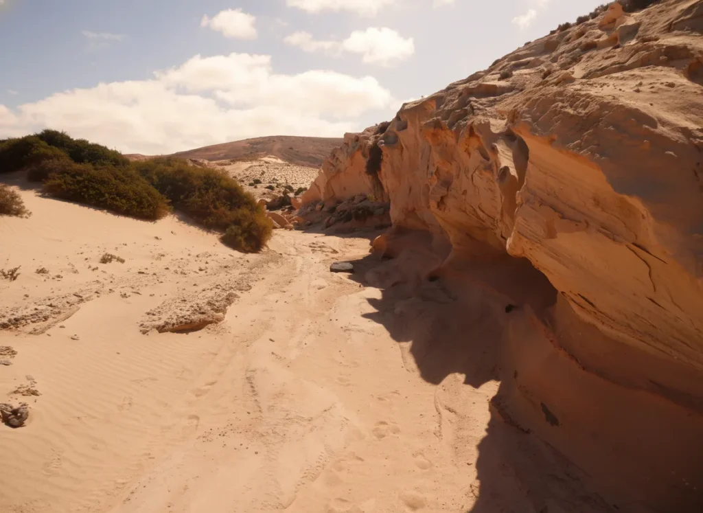Formación rocosa de arenisca naranja y marrón con erosión geológica en paisaje desértico, rodeada de arena clara y vegetación arbustiva bajo cielo nublado