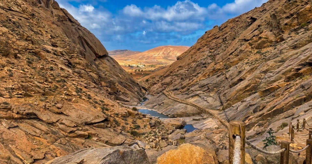Vista panorámica del Barranco de las Peñitas en Fuerteventura, mostrando un paisaje rocoso de tonos áridos, un pequeño sendero con barandilla de madera en primer plano, charcos de agua en el fondo del cañón y montañas al fondo bajo un cielo parcialmente nublado.