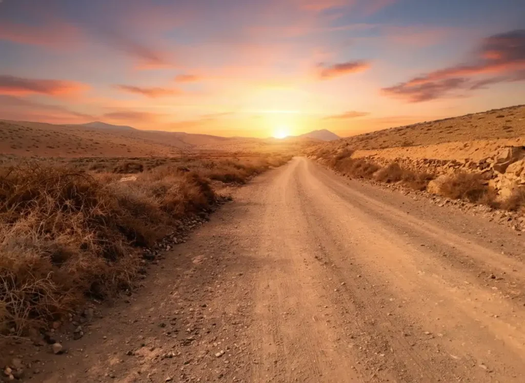 Carretera de tierra sin asfaltar atravesando un paisaje desértico árido con vegetación escasa, iluminada por el atardecer dorado con montañas visibles en el horizonte