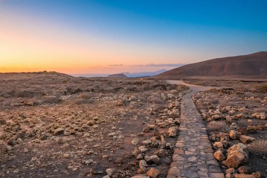 Sendero empedrado atravesando un paisaje desértico árido con rocas dispersas, colinas rocosas a ambos lados y un río o arroyo al fondo, iluminado por la luz dorada del atardecer con cielo azul gradual