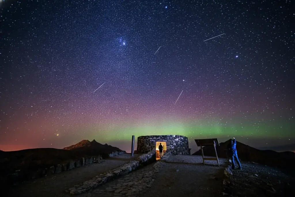 Persona observando la aurora boreal verde y rosa iluminando el cielo nocturno estrellado, con varios meteoros visibles y ruinas de piedra antigua en primer plano