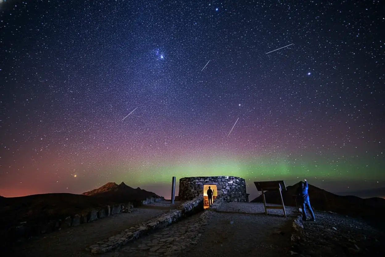 Persona observando la aurora boreal verde y rosa iluminando el cielo nocturno estrellado, con varios meteoros visibles y ruinas de piedra antigua en primer plano