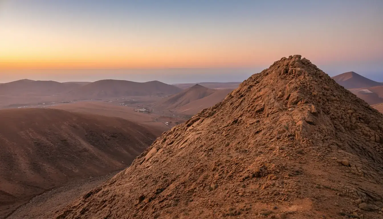 Vista panorámica de un paisaje volcánico desértico durante la hora dorada del atardecer, con montañas de arena y roca volcánica en primer plano, múltiples colinas volcánicas en el fondo y un cielo gradiente de tonos naranjas y rosas