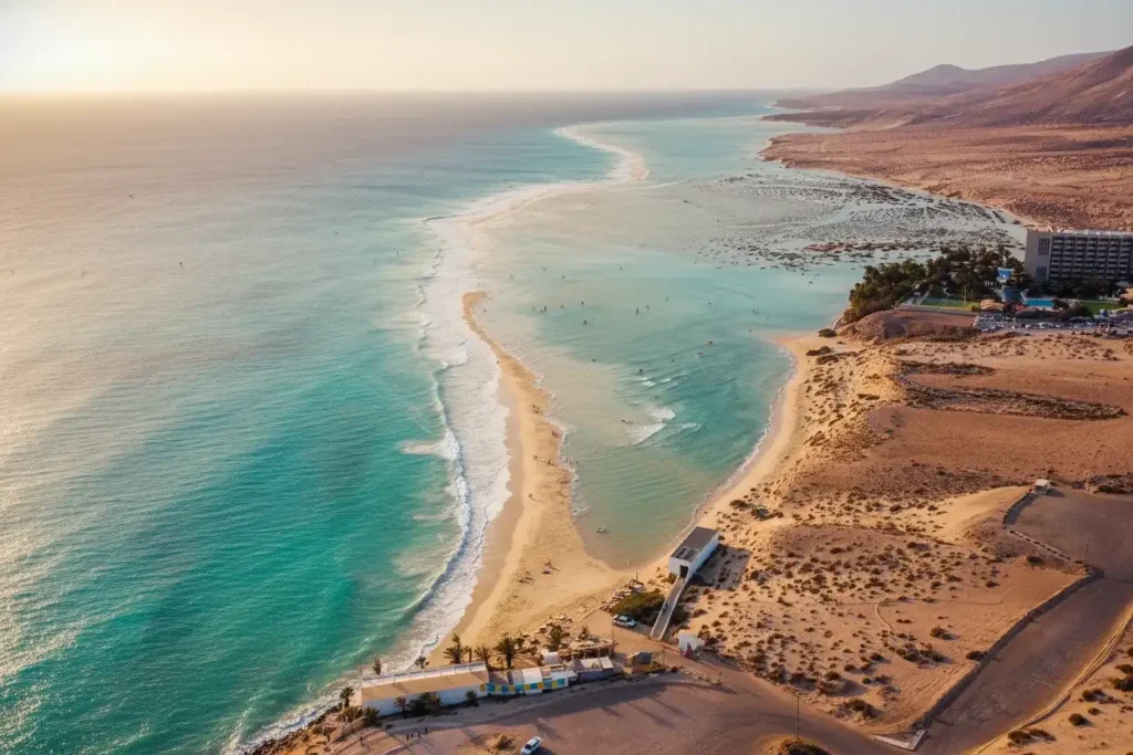 Vista aérea de una hermosa playa con aguas turquesas cristalinas, arena dorada, olas blancas, resort hotelero y paisaje desértico montañoso al atardecer