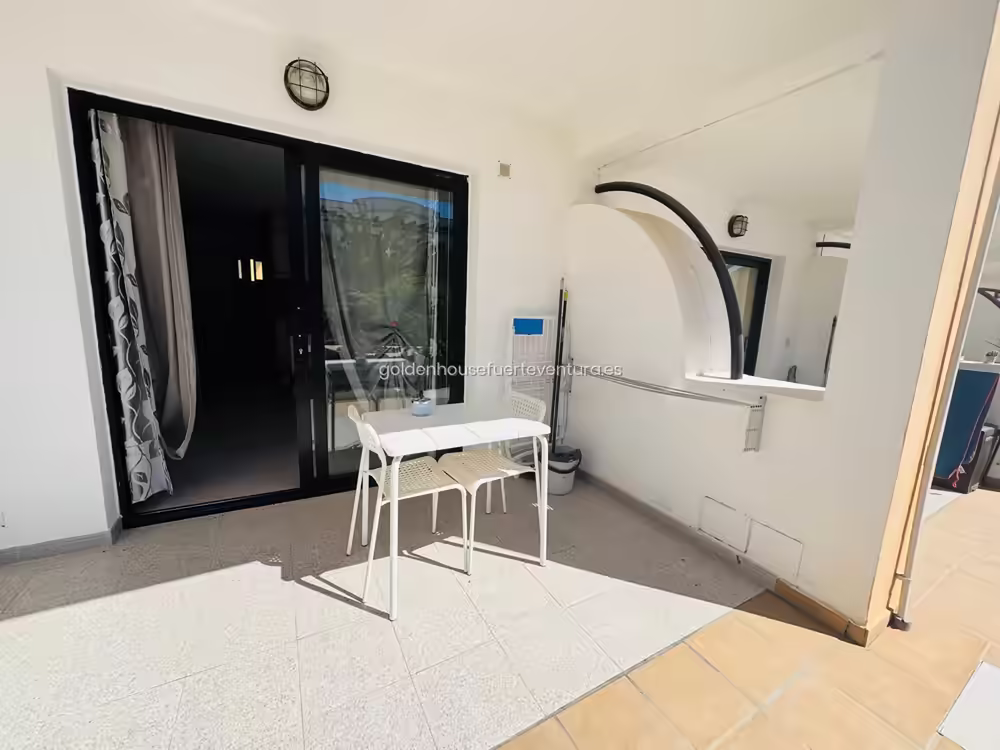 Terraza exterior con mesa blanca, dos sillas y acceso por puerta corredera acristalada.