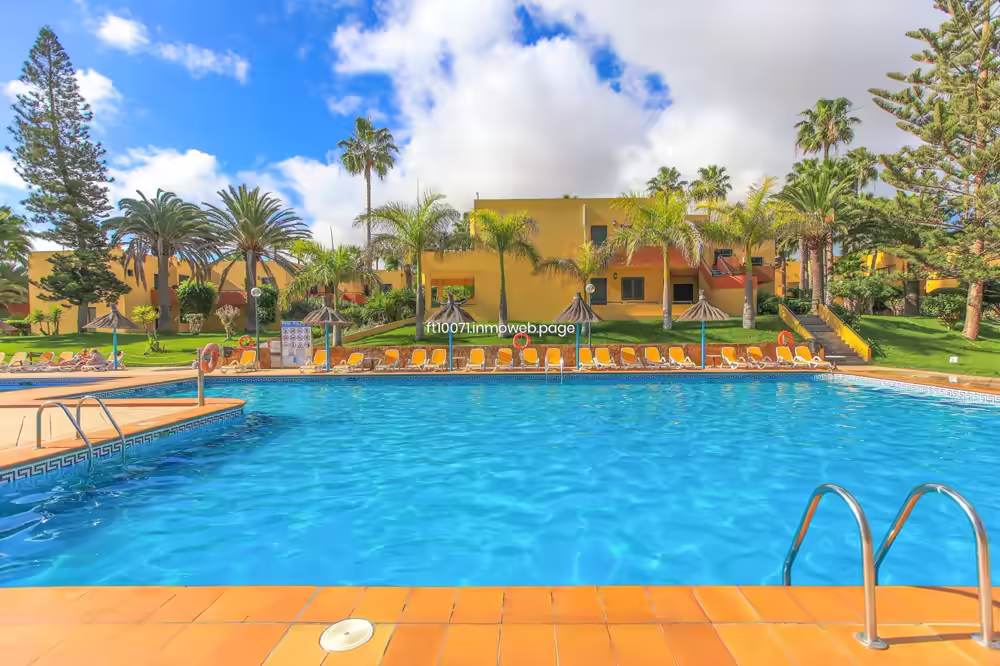Piscina comunitaria con hamacas amarillas y sombrillas bajo el cielo azul de Canarias.