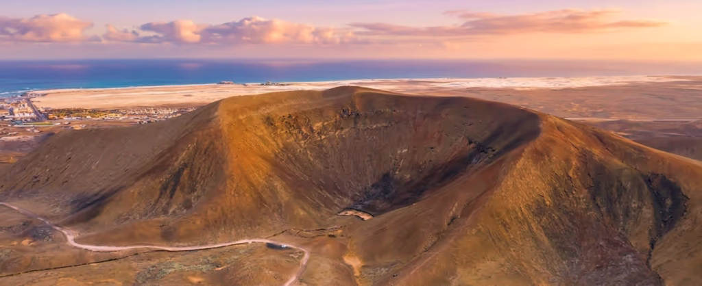 Vista aérea del cráter del volcán de Bayuyo en Fuerteventura durante el atardecer, mostrando tonos áridos y rojizos con el océano Atlántico y la costa al fondo bajo un cielo despejado con nubes ligeras.