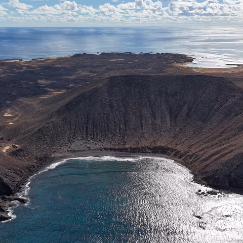 Vista aérea del cráter principal del volcán del Islote de Lobos, mostrando su apertura hacia el mar y la playa de arena negra interior.