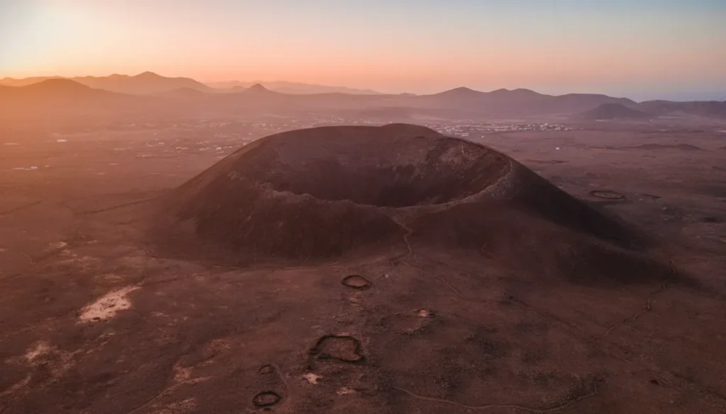 Vista aérea de un cráter volcánico con borde elevado y depresión central, rodeado de terreno árido y montañas distantes durante la puesta de sol