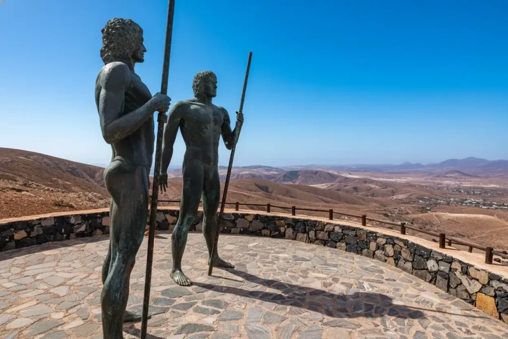 Dos estatuas de bronce de guerreros indígenas desnudos sosteniendo lanzas, ubicadas en un mirador con vista panorámica a un paisaje árido y montañoso