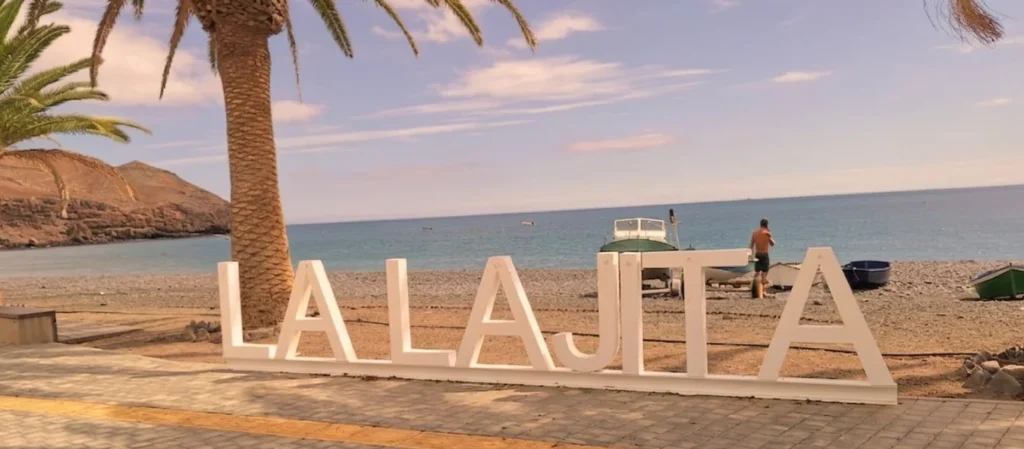 Gran letrero blanco en una playa tropical con palmeras, arena negra, mar turquesa y embarcaciones. Paisaje costero idílico con acantilados al fondo y cielo despejado