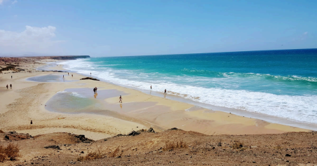 Vista panorámica de Piedra Playa con charcos en la arena por la marea, oleaje blanco y mar turquesa bajo un cielo despejado.