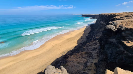 Vista en perspectiva desde un alto acantilado de roca hacia la playa de la Escalera con olas turquesas y arena dorada.