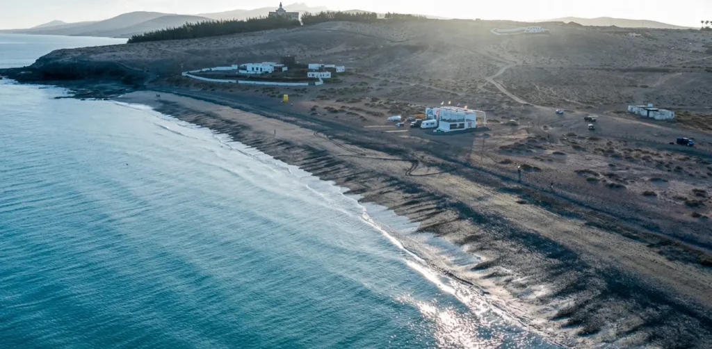 Vista aérea de una playa de arena negra volcánica con aguas turquesas, casas blancas dispersas en el terreno árido, montañas y vegetación de coníferas
