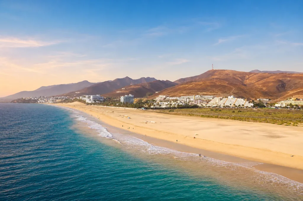Vista panorámica de una extensa playa de arena dorada con aguas turquesas, flanqueada por complejos hoteleros modernos y montañas áridas de color ocre al fondo bajo un cielo azul claro