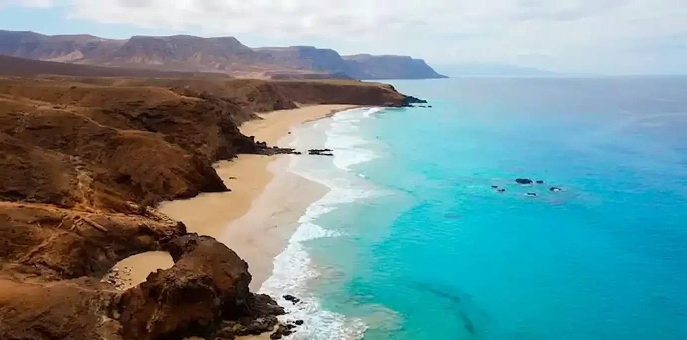Vista aérea de una playa virgen con arena blanca, acantilados rocosos marrones y aguas turquesas cristalinas, con montañas de meseta al fondo