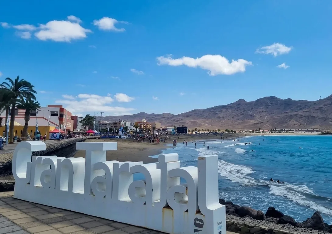 Letrero blanco en una playa tropical con olas turquesas, palmeras, montañas áridas de fondo y turistas disfrutando del mar en un día soleado