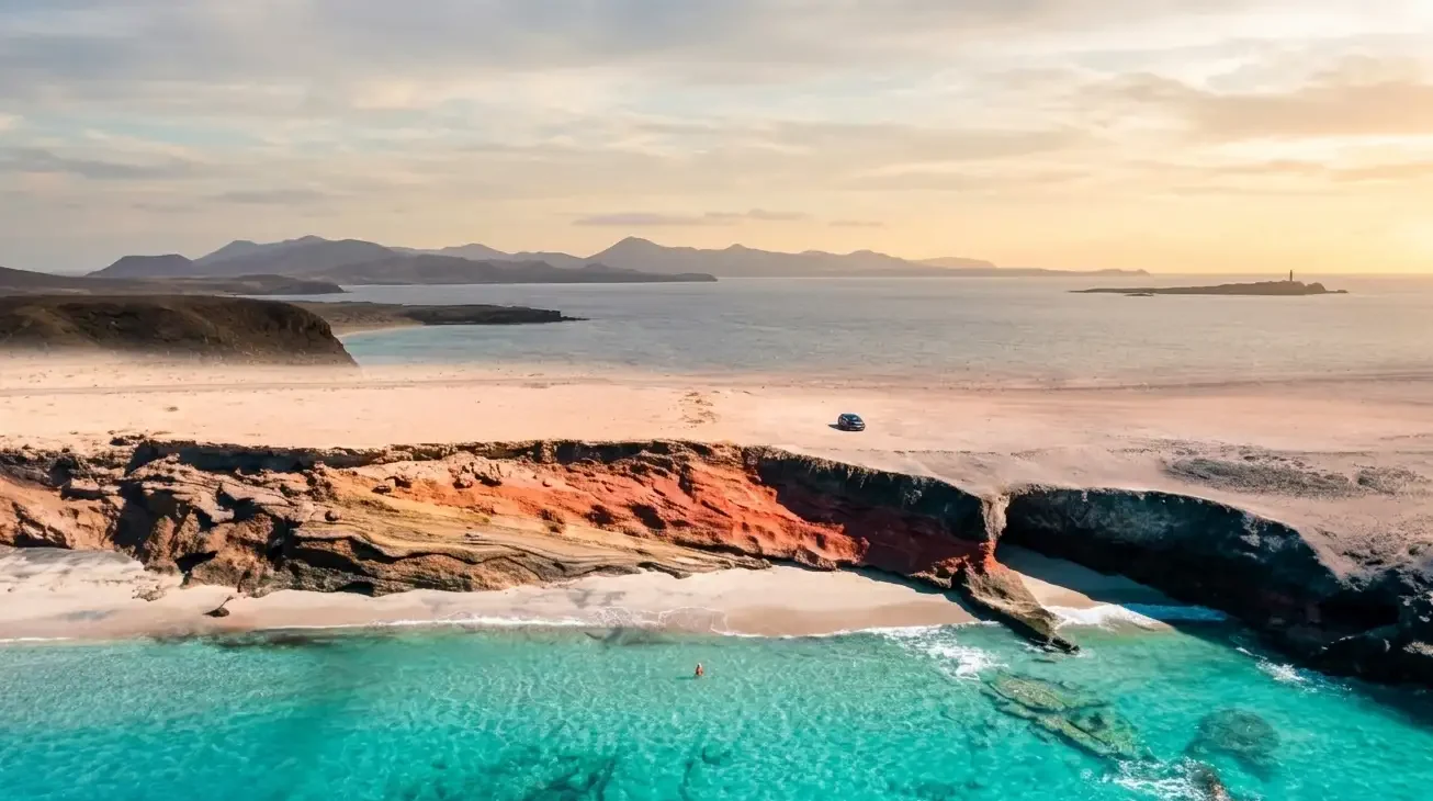 Vista aérea de una playa virgen con aguas turquesas cristalinas, arena blanca, acantilados de roca roja y naranja, pequeño vehículo en la orilla, islas montañosas al fondo y cielo dorado al atardecer