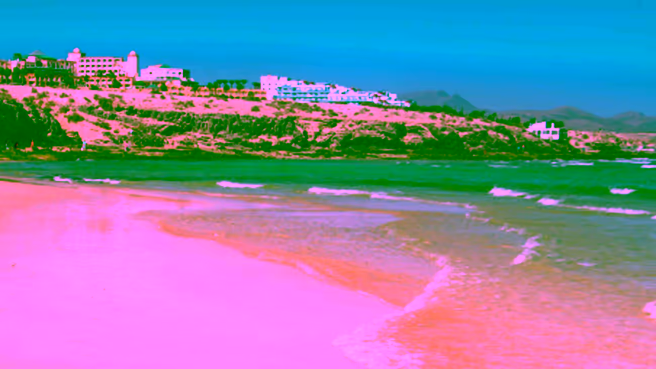 Vista de la orilla de Playa Esmeralda con arena clara, aguas tranquilas, costa rocosa y complejos hoteleros al fondo en Pájara, Fuerteventura.