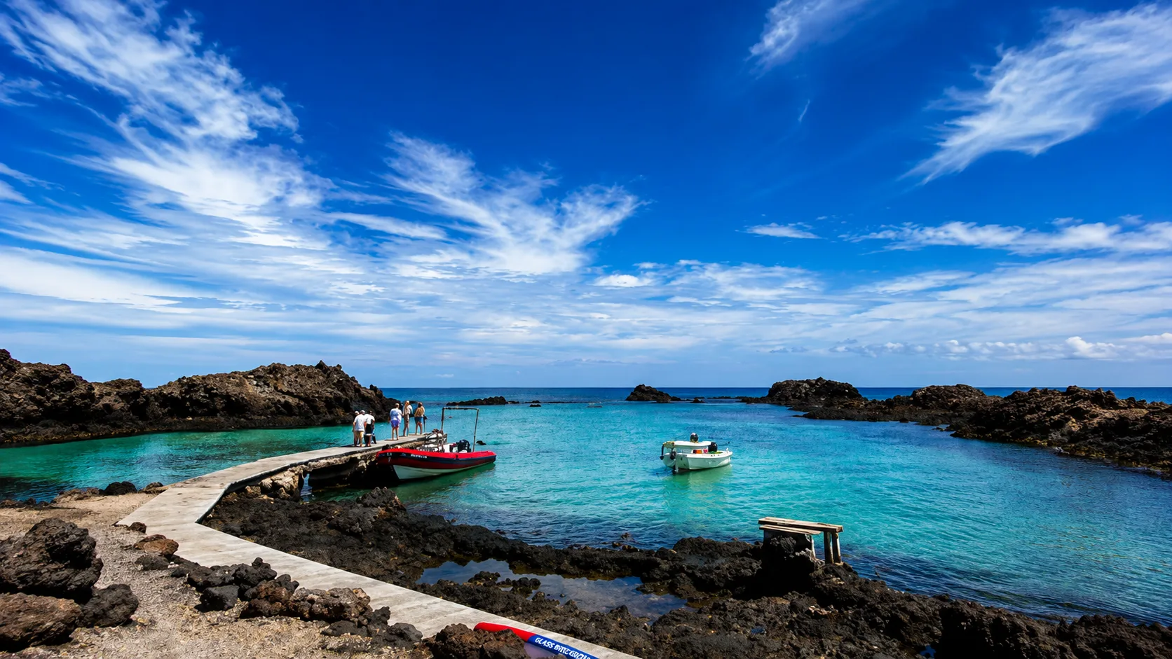 Embarcadero de la Isla de Lobos en Fuerteventura con aguas turquesas, pequeñas embarcaciones y paisaje volcánico bajo un cielo azul.