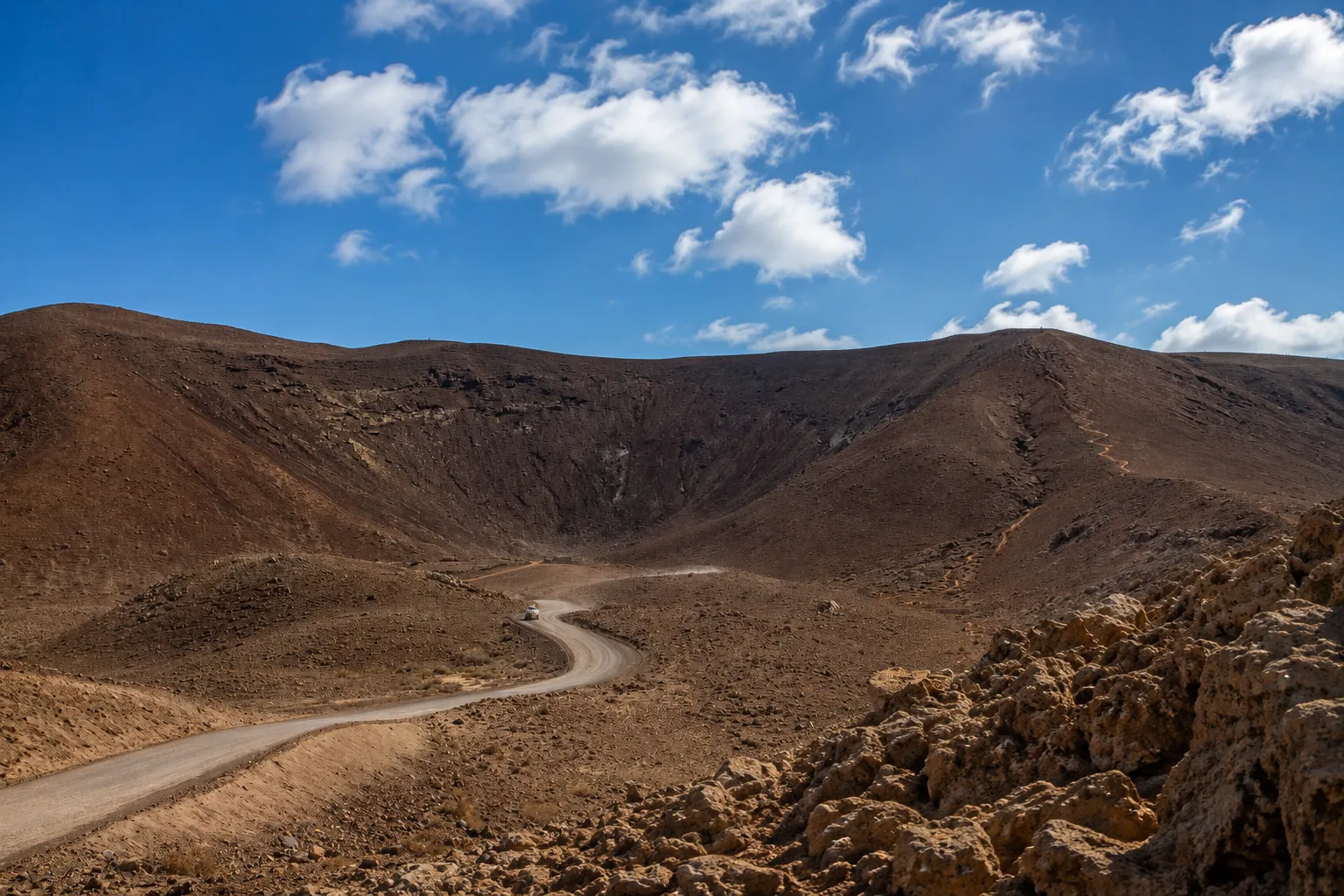 Camino de tierra serpenteante que atraviesa laderas de conos volcánicos y terrenos áridos erosionados en Fuerteventura bajo un cielo azul con nubes.