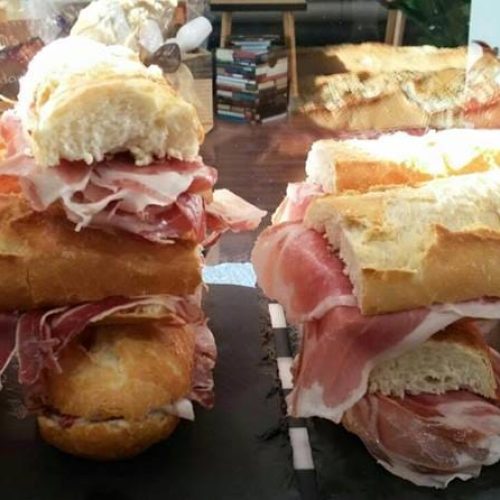Dos grandes bocadillos de pan de barra rellenos generosamente con múltiples capas de jamón serrano, expuestos en el mostrador de un bar.