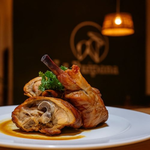 Ración de cabrito asado majorero servida sobre salsa en plato blanco, con fondo del restaurante La Puipana desenfocado.