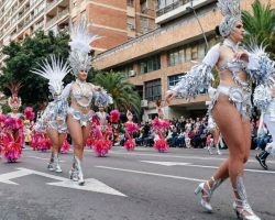 Bailarinas con trajes plateados y plumas desfilan por una avenida, con público a ambos lados y palmeras al fondo.