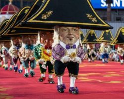 Personajes con cabezas gigantes y sombreros negros desfilan en fila sobre una alfombra roja, con público alrededor.
