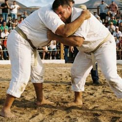 Dos luchadores con ropa blanca se agarran en un terrero de arena, con gradas llenas de público al fondo.