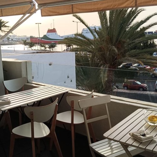 Terraza del restaurante Zielo Terraza al atardecer, con mesas y sillas blancas, y vistas al puerto con un ferry y palmeras.