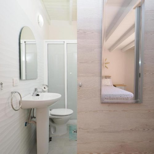 Vista de aseo con lavabo blanco y espejo que refleja un dormitorio con techo de madera.