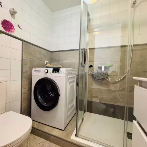 Baño con azulejos blancos, ducha de vidrio y lavadora de carga frontal.