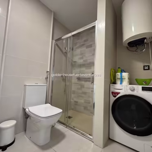 Baño con ducha de cristal, inodoro blanco y lavadora de carga frontal.
