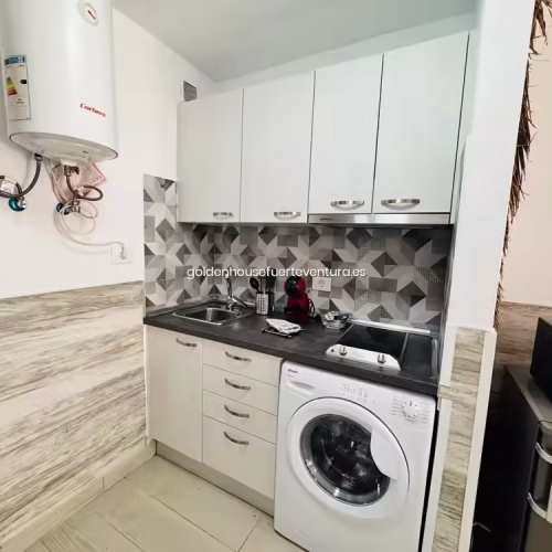 Cocina lineal con armarios blancos, vitrocerámica de dos fuegos y lavadora de carga frontal.