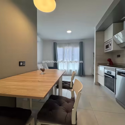 Mesa de madera con sillas blancas frente a cocina lineal equipada.