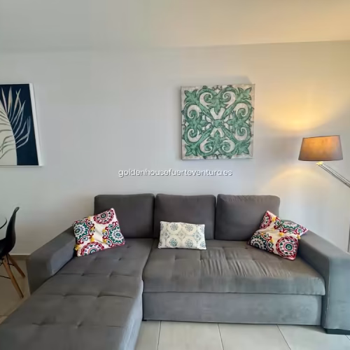 Sofá chaise longue gris con cojines estampados y cuadros decorativos en la pared.