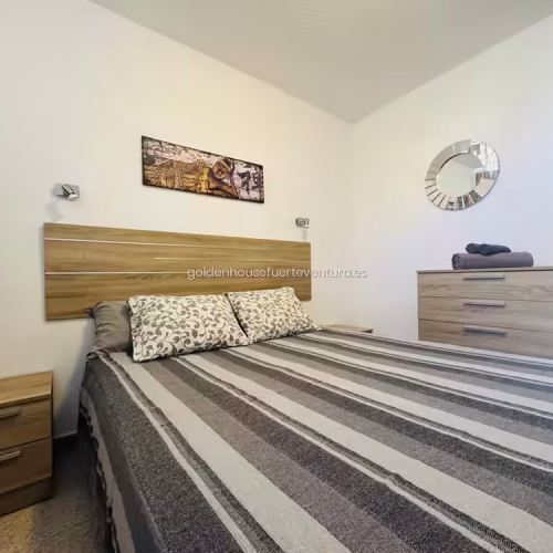 Vista lateral de la cama doble con almohadas estampadas y cómoda de madera.
