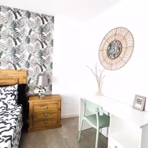 Mesita de noche de madera con lámpara plateada y detalle de pared decorada.