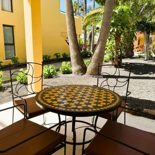Primer plano de mesa de terraza con sillas y vistas a palmeras y zonas comunes del complejo.