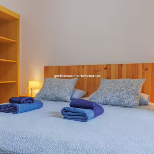 Cama de matrimonio con ropa de cama azul, cabecero de madera y armario abierto lateral.