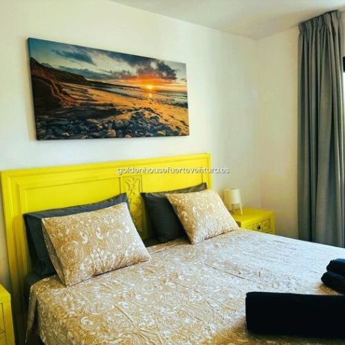 Cama doble con cabecero amarillo, cojines estampados y cuadro de puesta de sol sobre la cama.