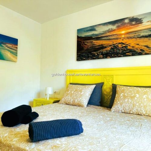 Dormitorio con cabecero amarillo, cama doble, cojines estampados y cuadros marinos.