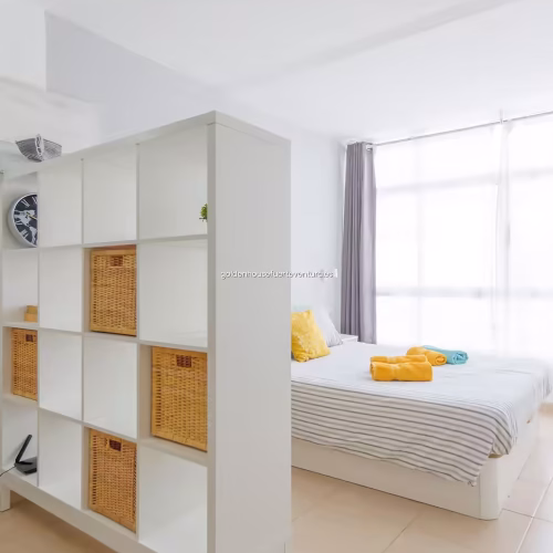 Cama de matrimonio junto a estantería blanca con cestas de mimbre en un espacio abierto