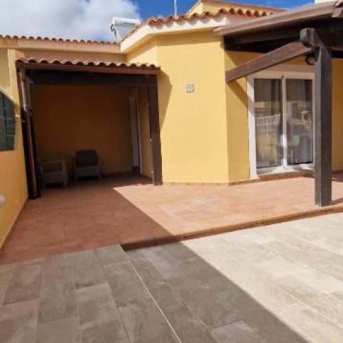 Fachada de color amarillo con porche de madera y paneles solares en el techo.