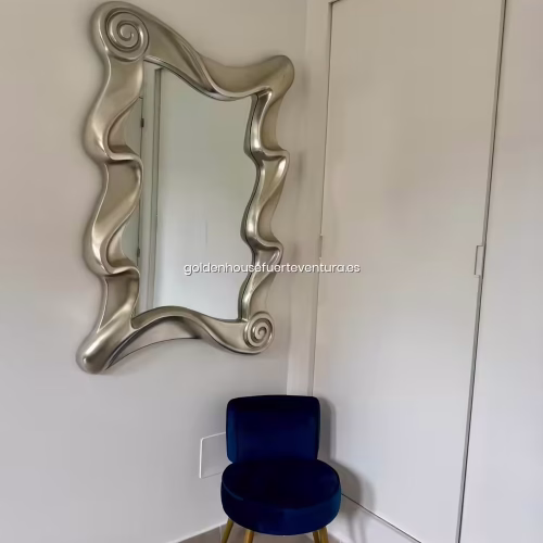 Espejo con marco plateado ondulado sobre silla de terciopelo azul en el dormitorio.