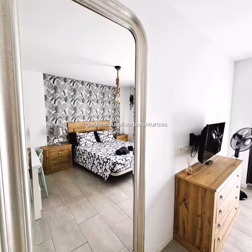 Espejo de cuerpo entero con marco plateado reflejando cama doble y cómoda de madera con TV.